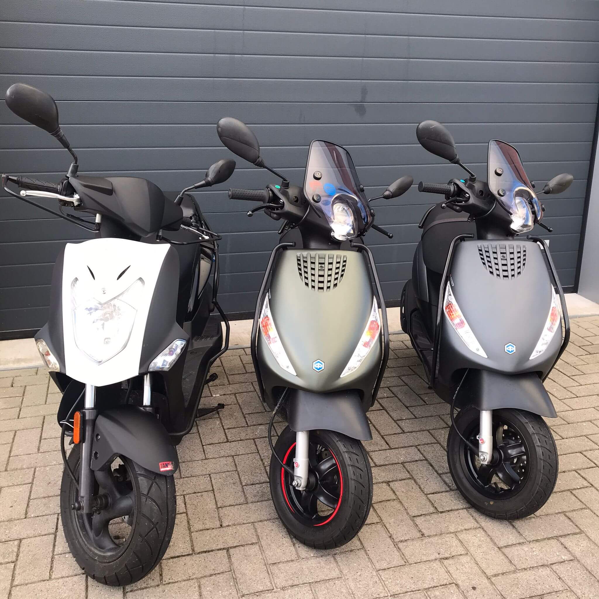 Drie scooters in neutrale kleuren, geparkeerd op een terrein met een grote grijze roldeur zichtbaar op de achtergrond